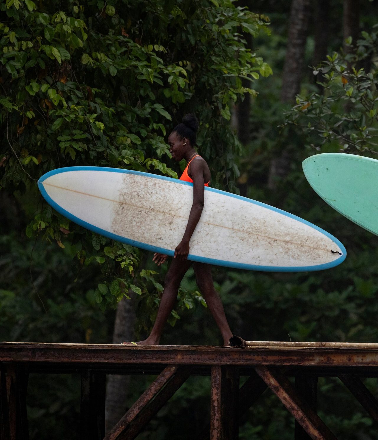 Femme à São Tomé marchant sur un pont en fer, avec une planche de surf à la main, avec de la végétation derrière