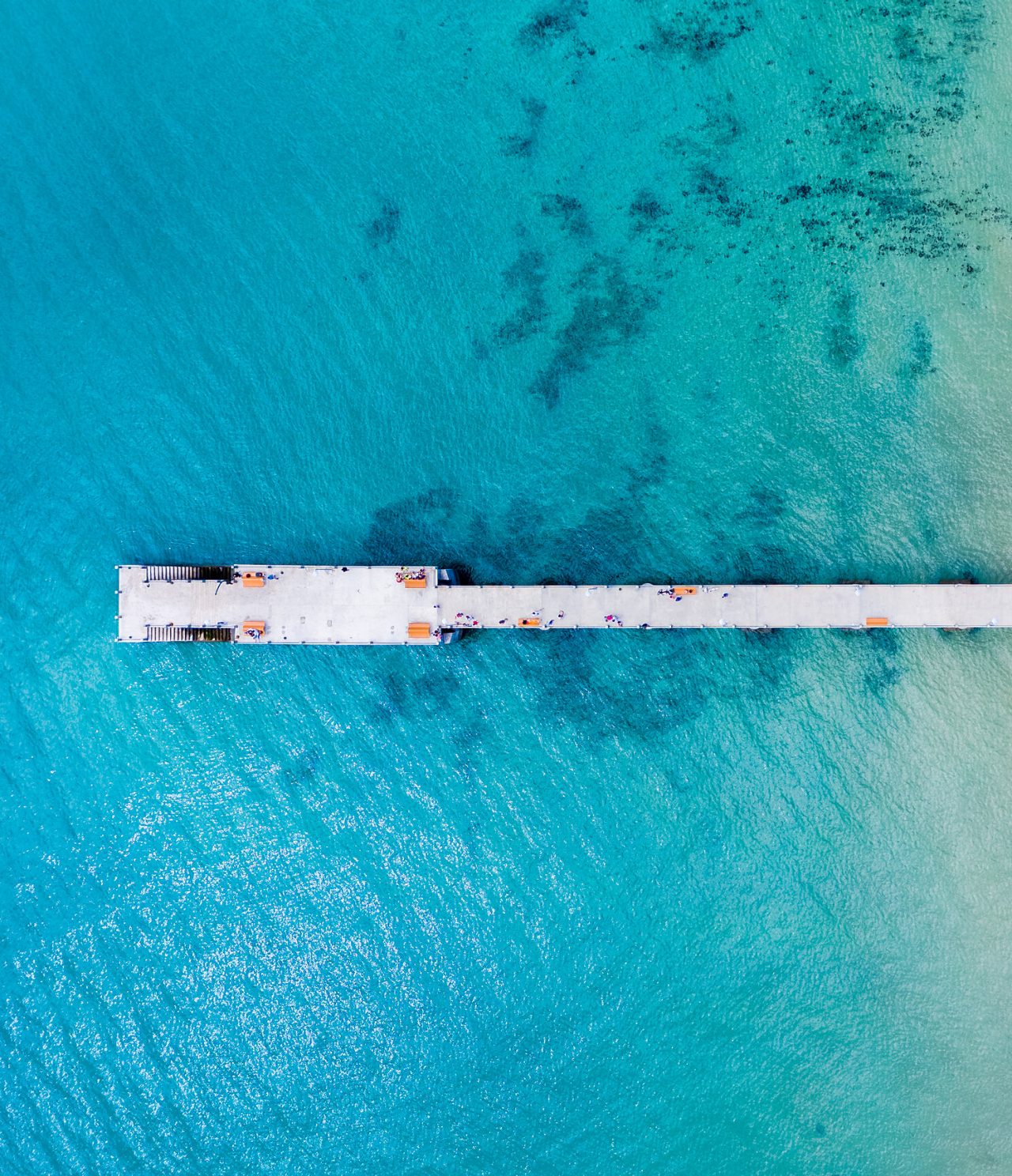 Vue aérienne d'un ponton en ciment, entouré de mer bleu turquoise, avec des escaliers pour descendre à l'eau