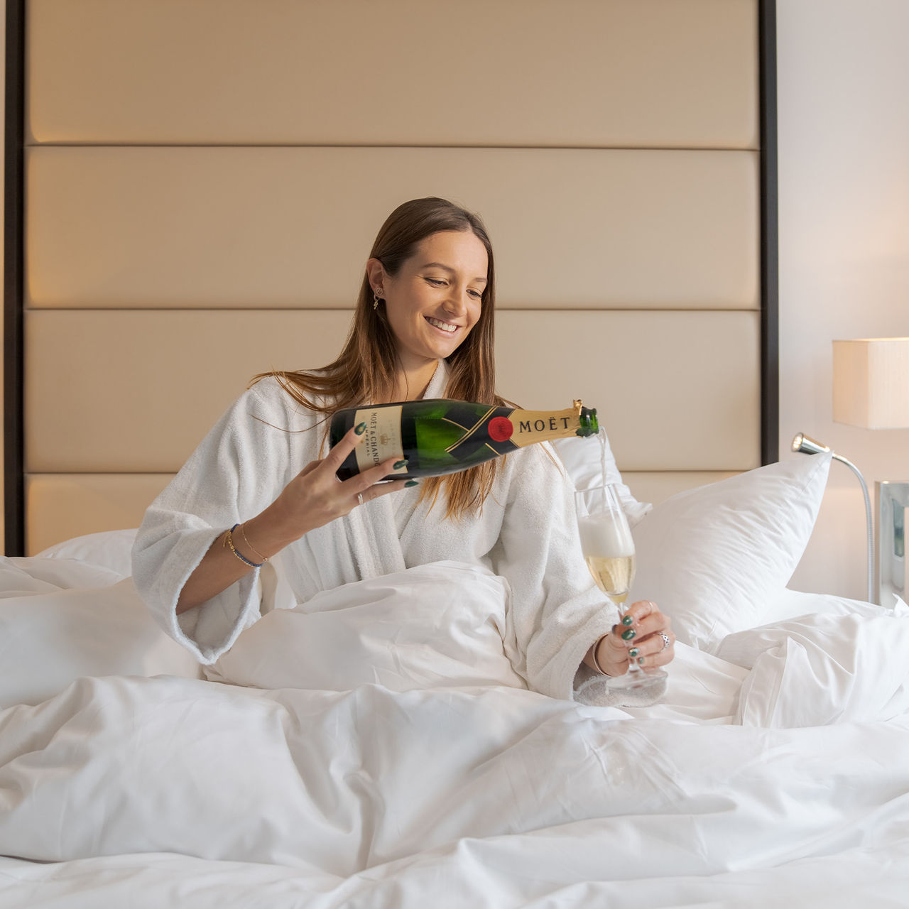 Fille souriante assise sur le lit avec un peignoir blanc et une bouteille de champagne