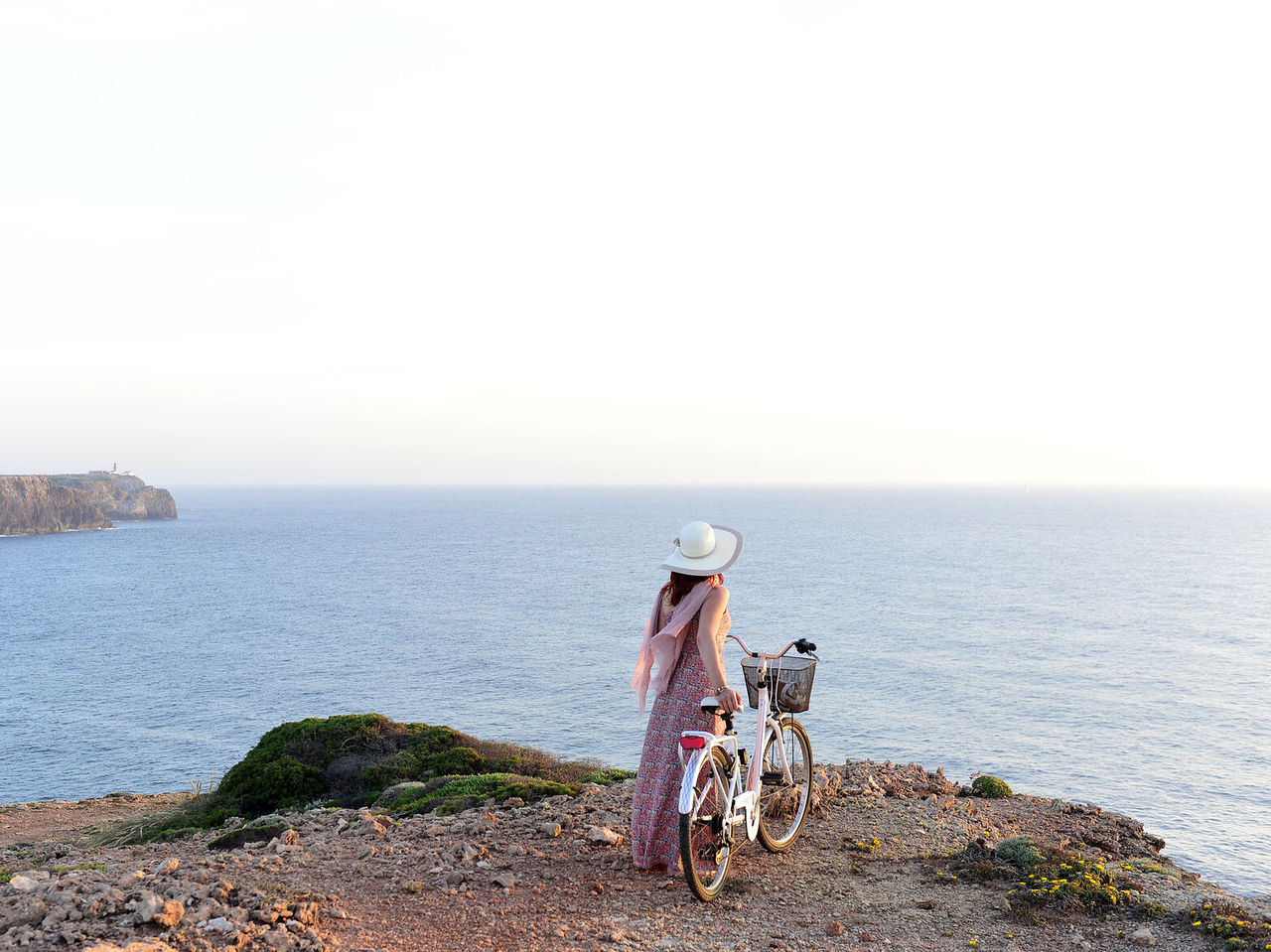 Jonge vrouw met een hoed en een fiets in de hand, genietend van het uitzicht op de zee vanaf een klif in de Algarve