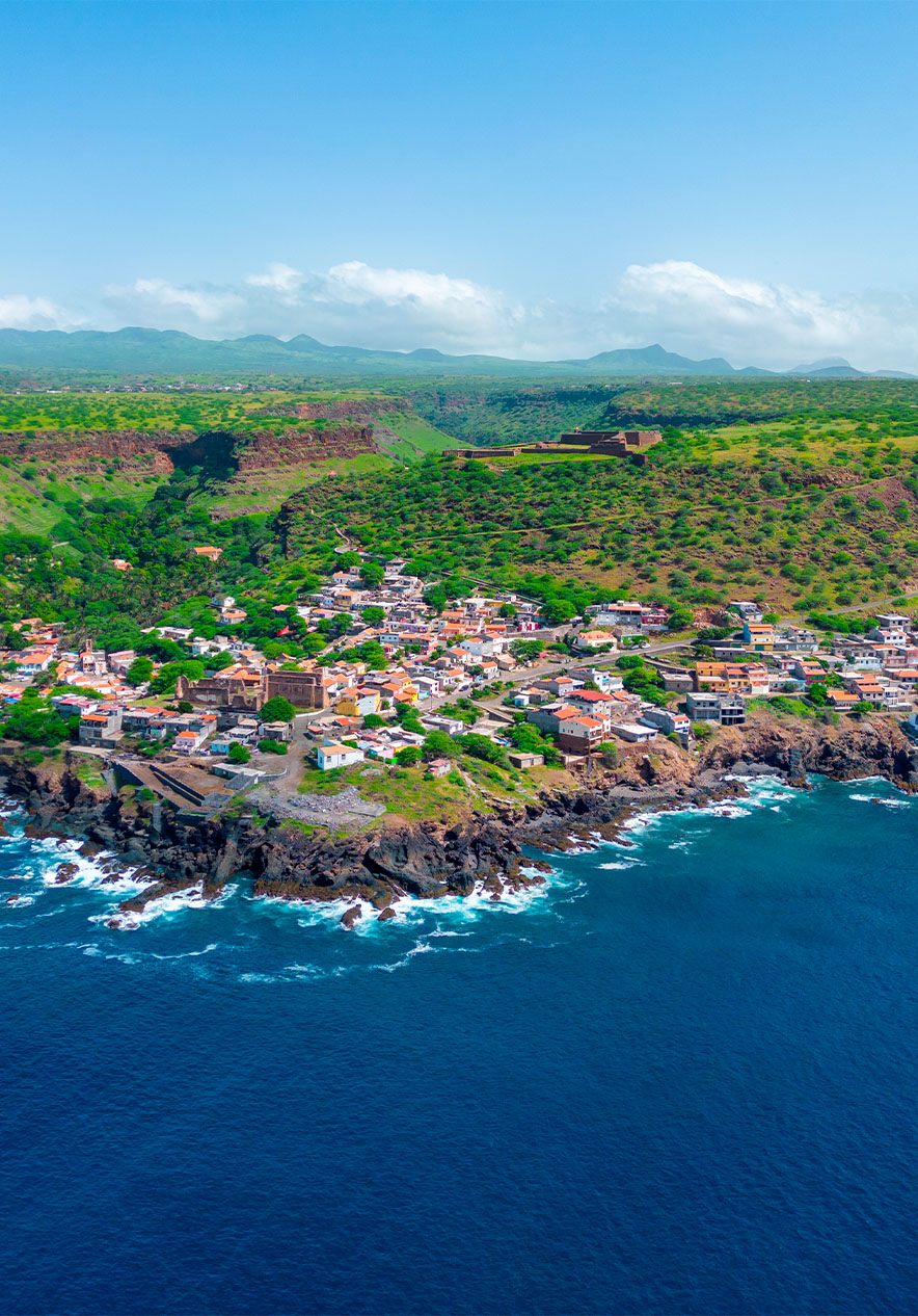vista da Cidade Velha, na ilha de Santiago, com o mar azul, várias casas, intensa vegetação e a zona montanhosa ao fundo