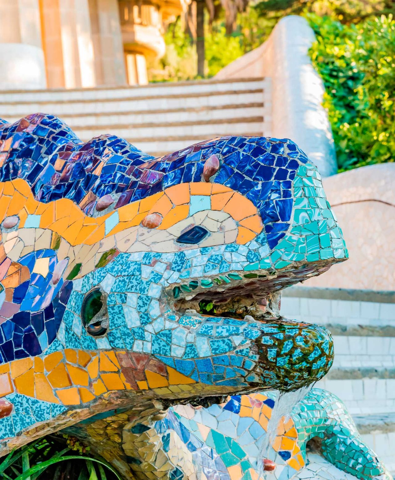 A icónica escultura de um dragão colorido, uma obra de Gaudí, no Parque Güell, em Barcelona, com escadas e vegetação no fundo