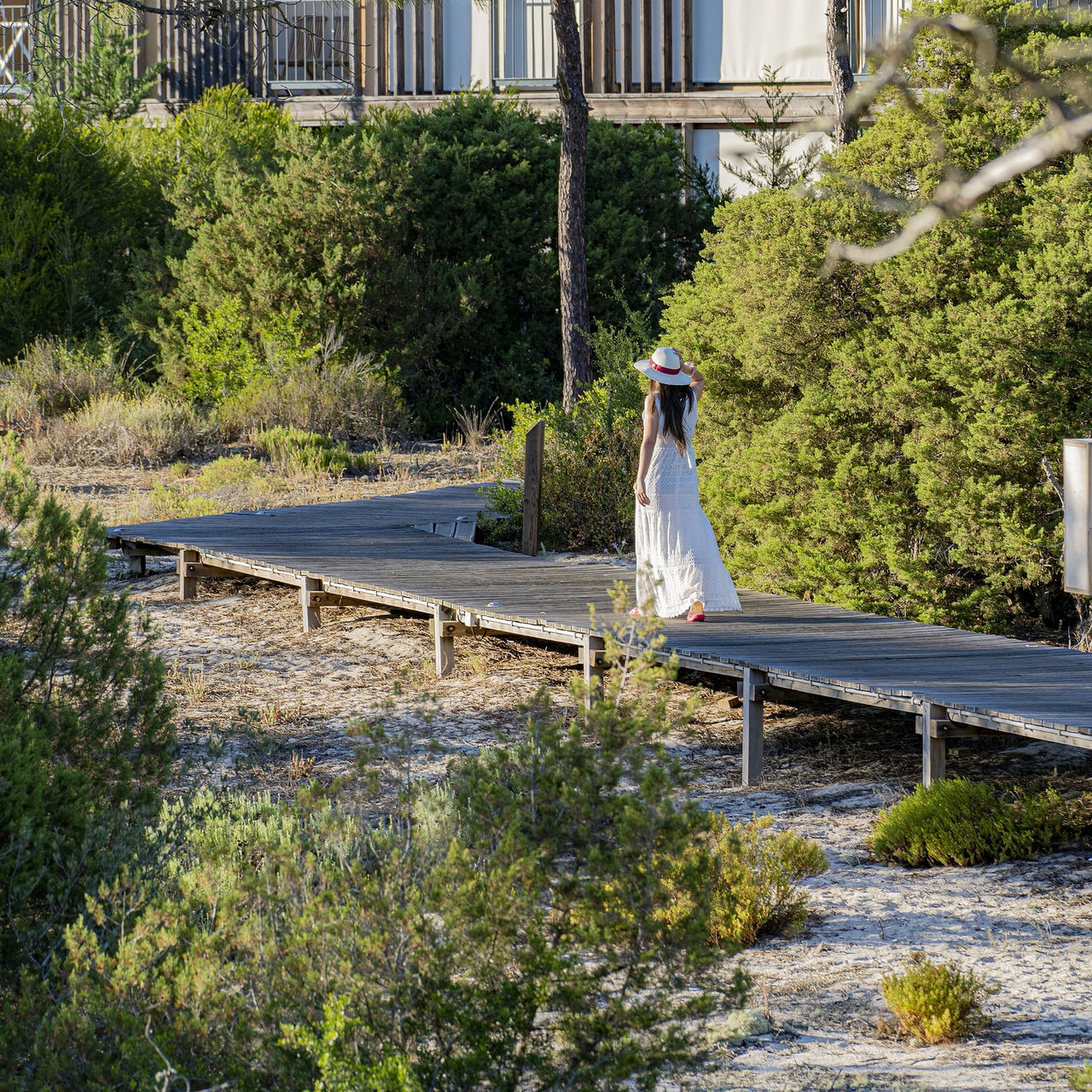 Passadiço de madeira no Pestana Tróia Eco Resort, onde uma mulher de vestido e chapéu branco passeia entre as villas