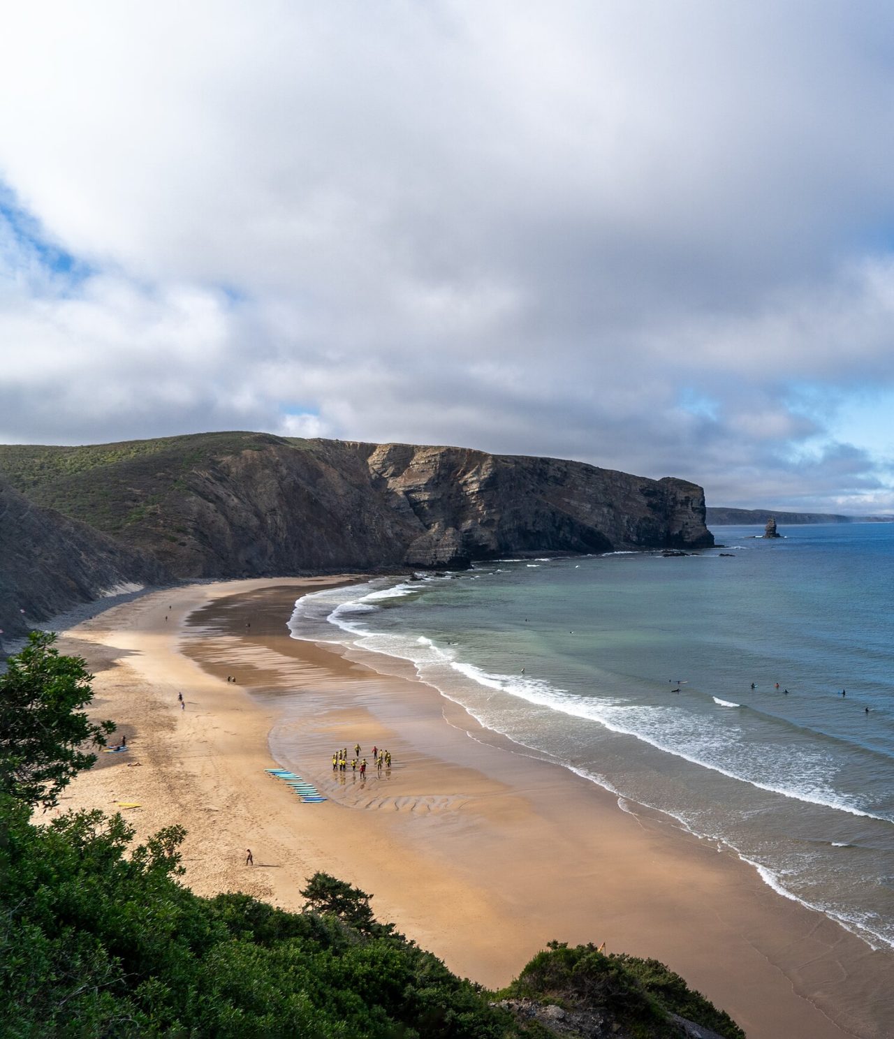 Vista panorâmica de uma praia extensa com falésias rochosas e surfistas nas ondas, na região do Algarve