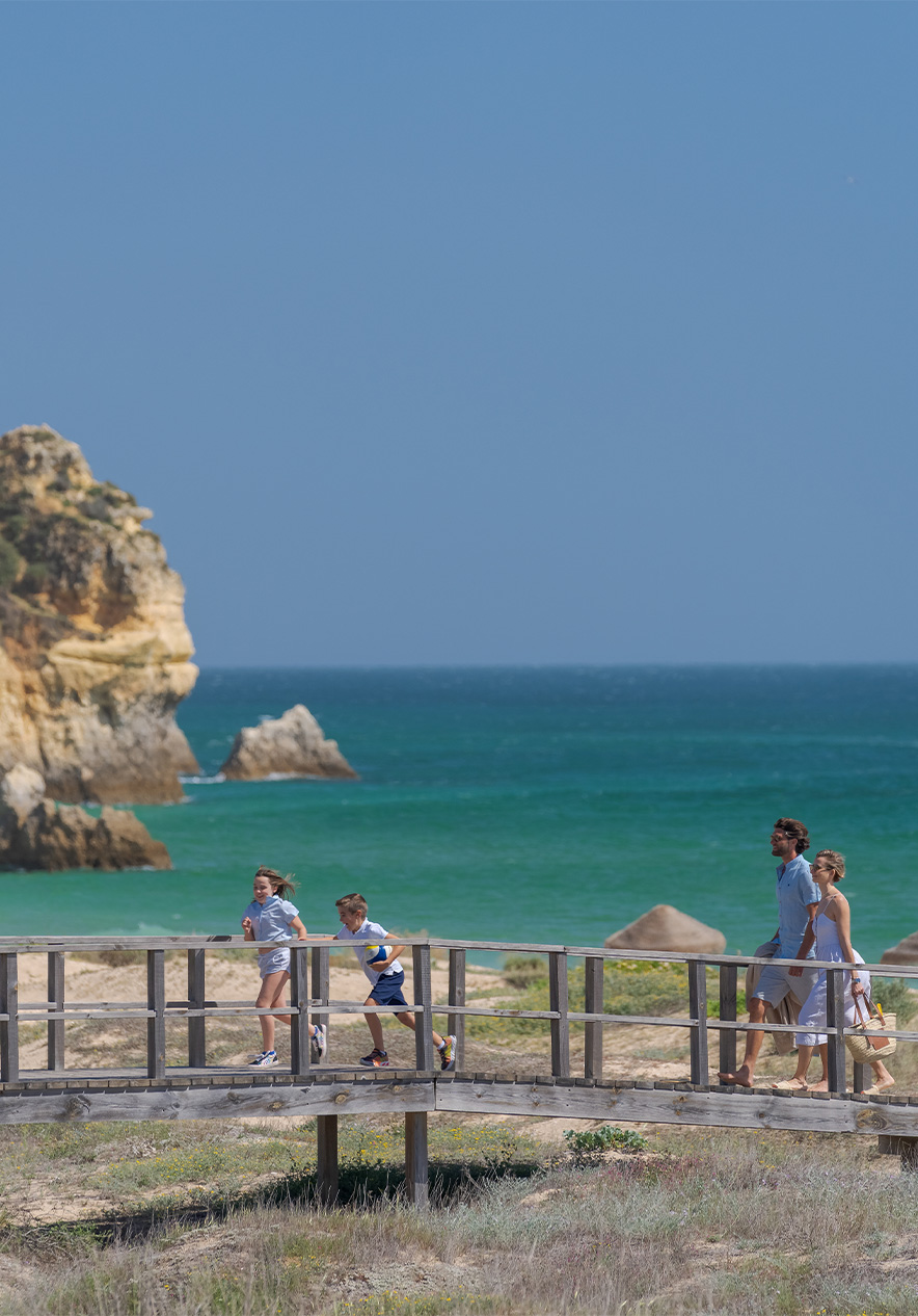 Passadiços de madeira das praias do Alvor, com o mar no fundo e duas crianças a correr com os pais atrás