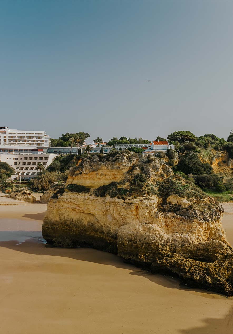 Praia do Alvor com rochas, areia, vegetação, a falésia e edifícios no fundo, com céu limpo