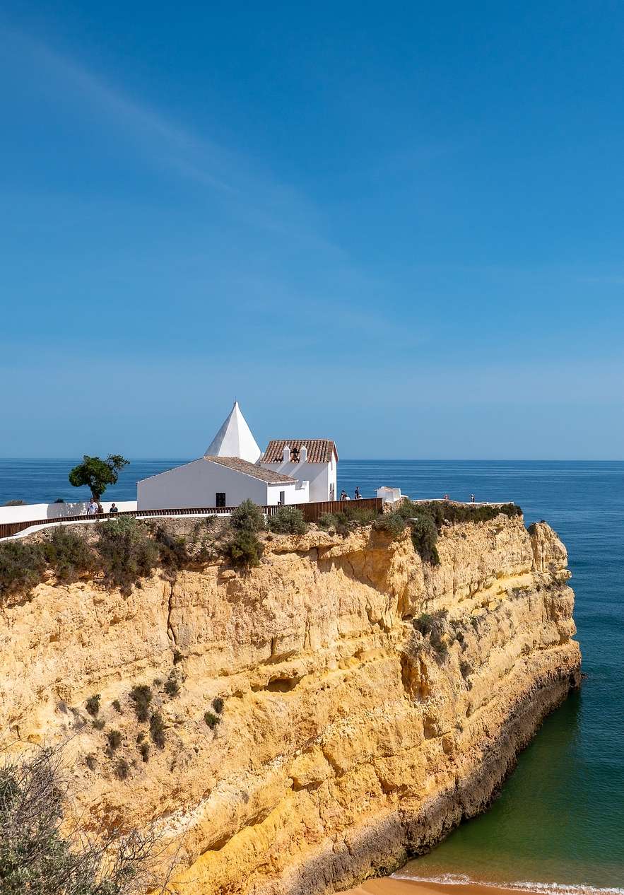 Vista sobre a falésia da praia senhora da rocha, com uma pequena casa branca cima da falésia, e um mar calmo ao fundo