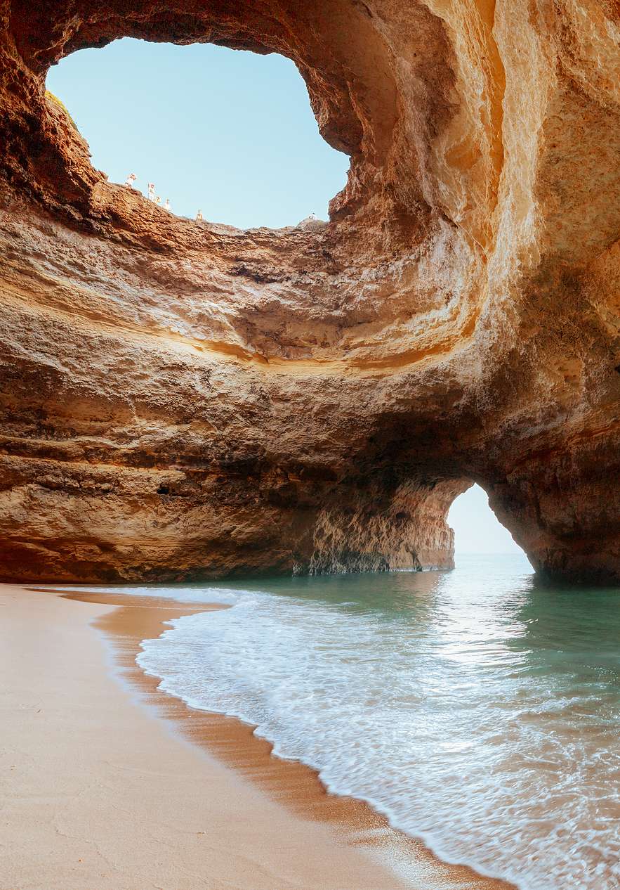 Caverna costeira no Algarve com um barco e turistas dentro, iluminada pela luz natural que entra por uma abertura no teto