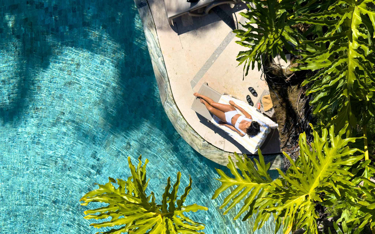 Senhora deitada numa espreguiçadeira à beira de uma piscina, cercada por palmeiras e vegetação tropical