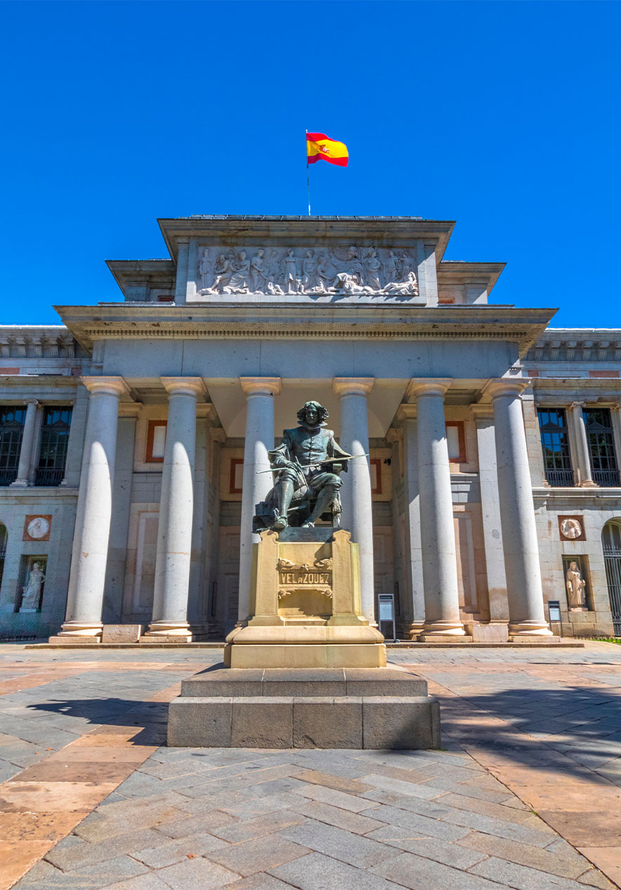 Facade of the Prado Museum with a statue at the entrance and the Spanish flag at the top of the building